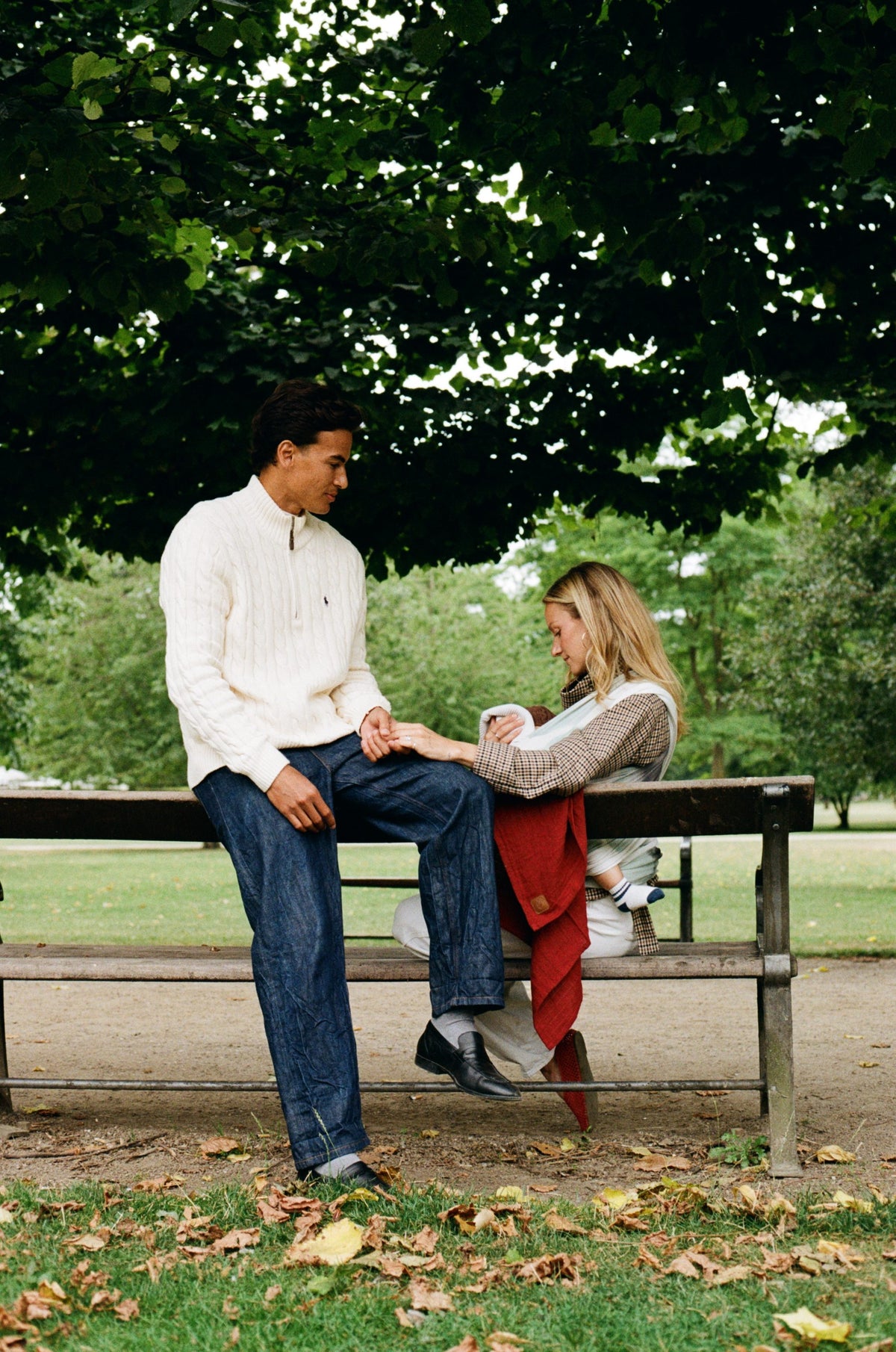 A couple on a park bench under the trees, the mother holding their baby in an Atala Red Cocoon Slingwrap.