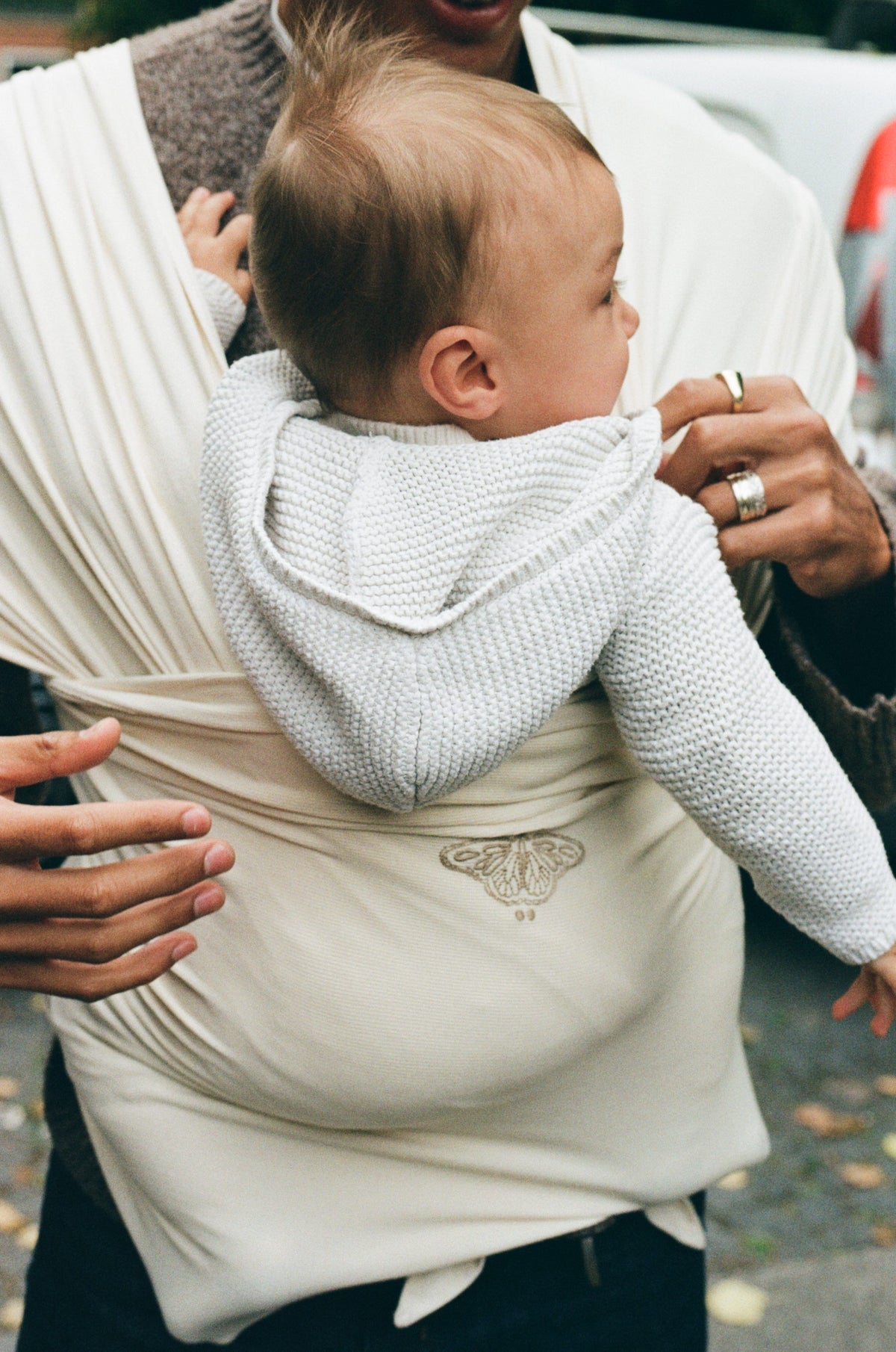A baby in a cream knit carried on a parent's chest in an Apollo Cream Cocoon Slingwrap, a hand resting on the wrap.