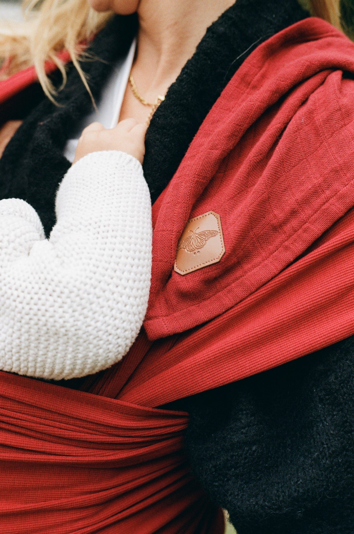 A baby in a white knit held close in an Atala Red Cocoon Slingwrap, the leather Salomë emblem on the wrap.