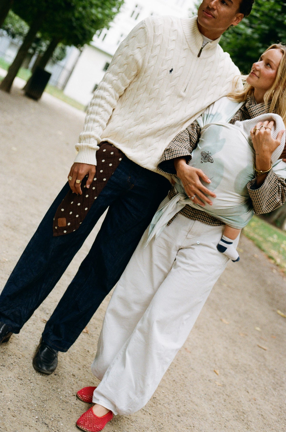 A couple walking through a Copenhagen park, the mother carrying their baby in a Pale Morpho Cocoon Slingwrap, the father holding a Pierrot Dot muslin cloth.